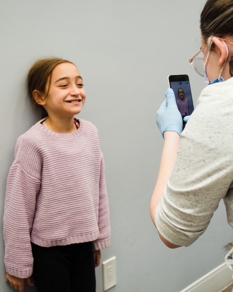 Hidden-smiles-early-orthodontic-treatment Child smiling during orthodontic consultation while staff member takes dental photos for treatment planning